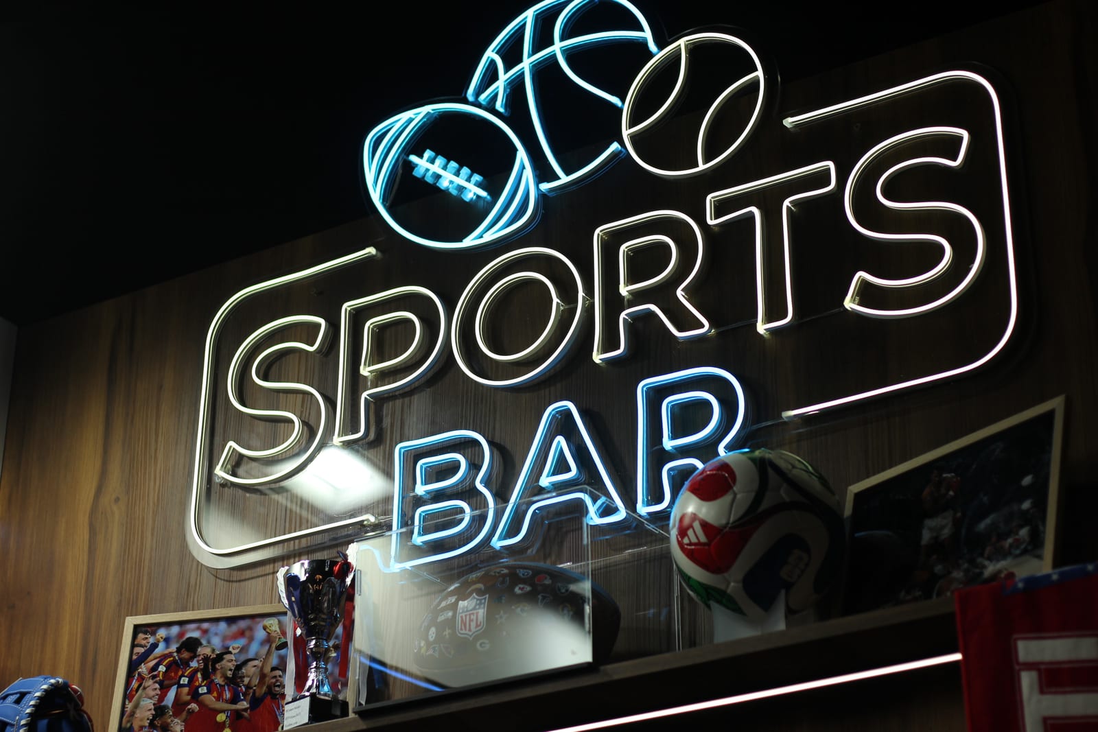 L'insegna al neon Sports Bar sopra il bancone dello stand 2Coders a IBC 2026, con una coppa, un casco NFL e un pallone da partita sulla mensola sottostante.